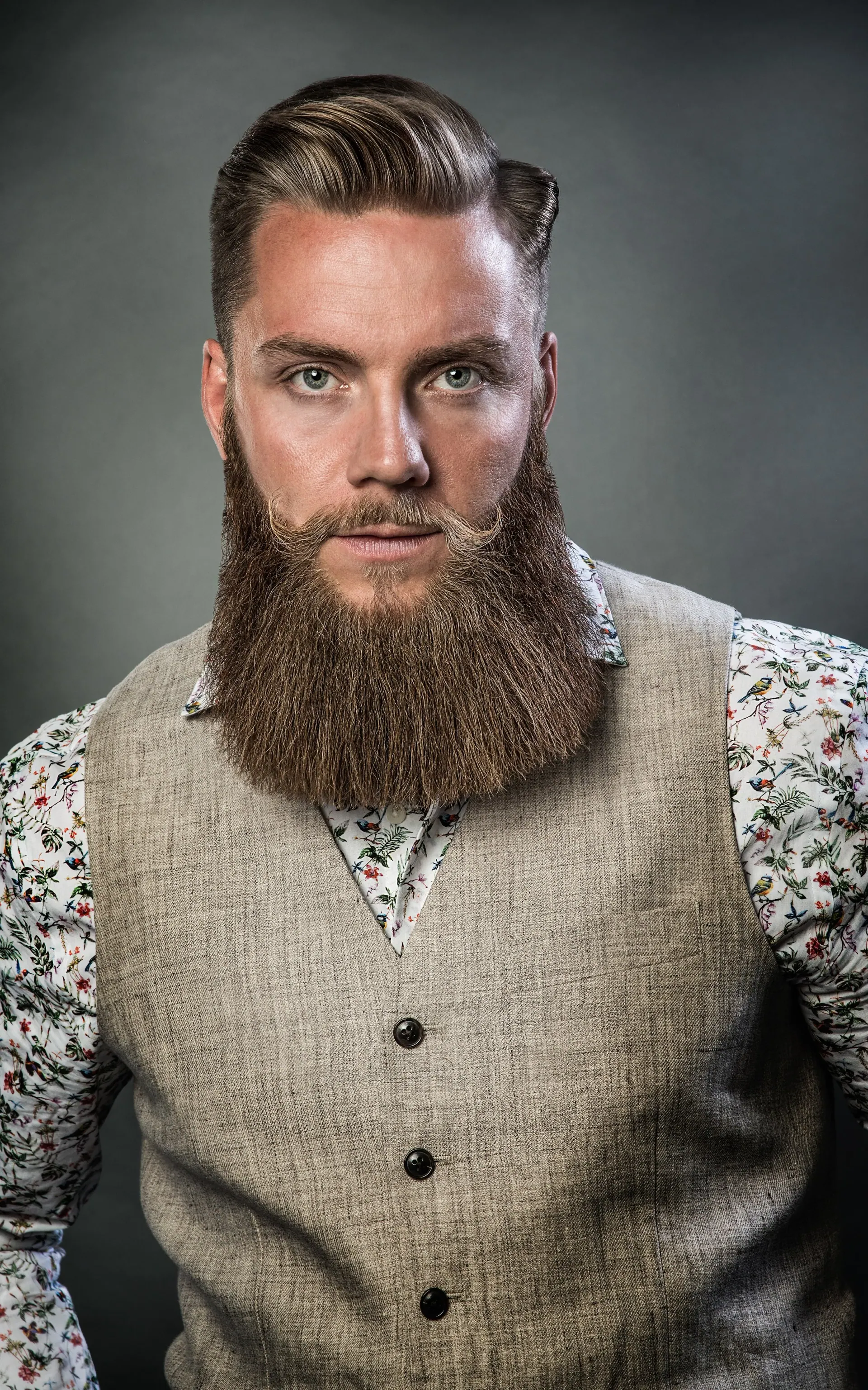Man med ett stort skägg och hårt stylat hår, bär en kavajväst över en blomstertröja med krage.