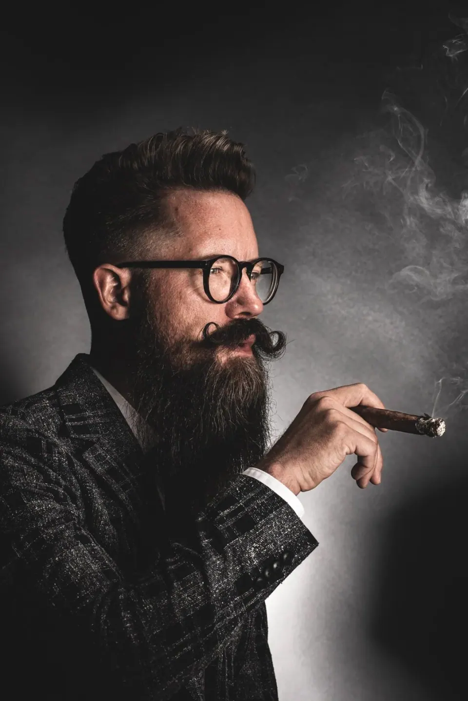 En man med helskägg och vaxat hår, bär glasögon och en svartvit rutig jacka, håller en cigarr.